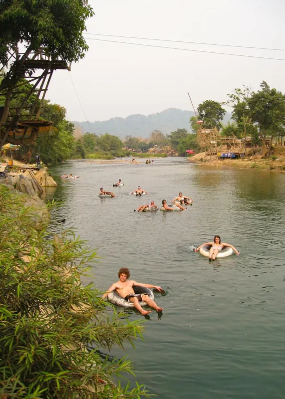 A serene river scene with people tubing, a wooden platform with onlookers, and a lush riverside landscape in Laos.