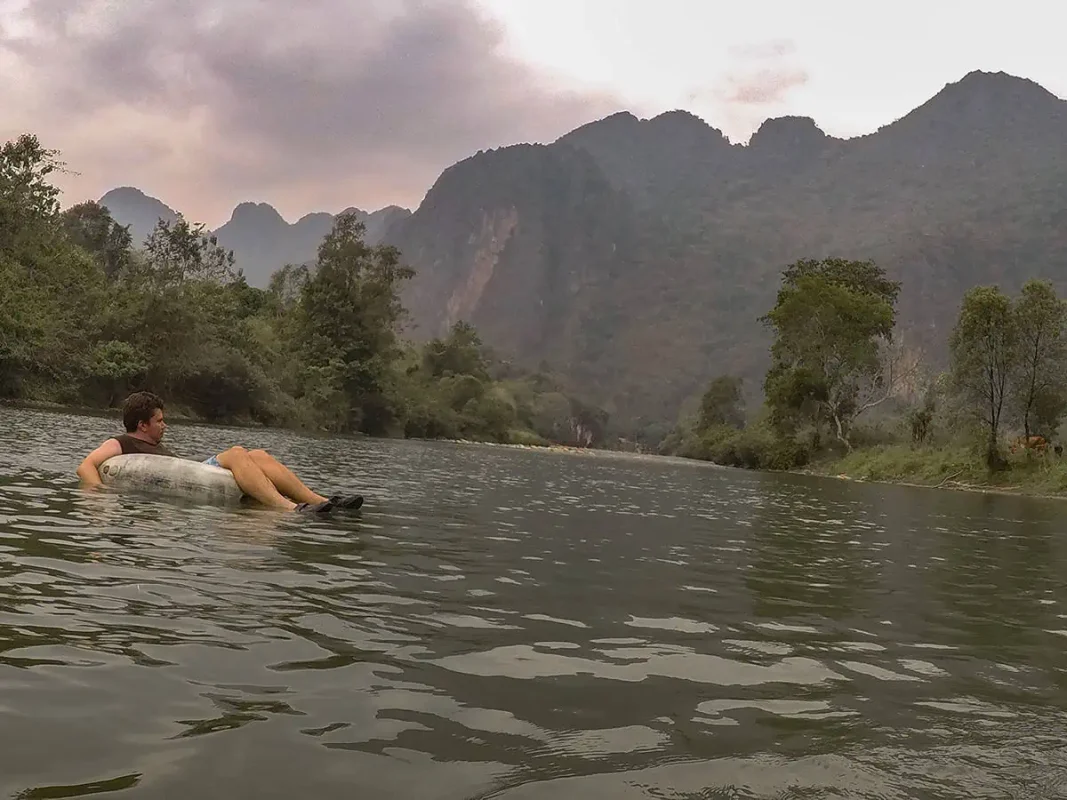 Tubing in Laos depicted by a man reclining in a tube on a river, surrounded by majestic mountains and a peaceful natural environment.