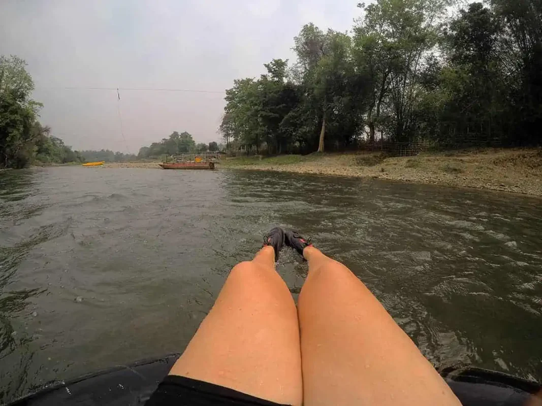A first-person view of a person's feet extended over a black inner tube, floating down a murky river with trees lining the shore and a yellow kayak in the distance, a typical scene of tubing in Vang Vieng.