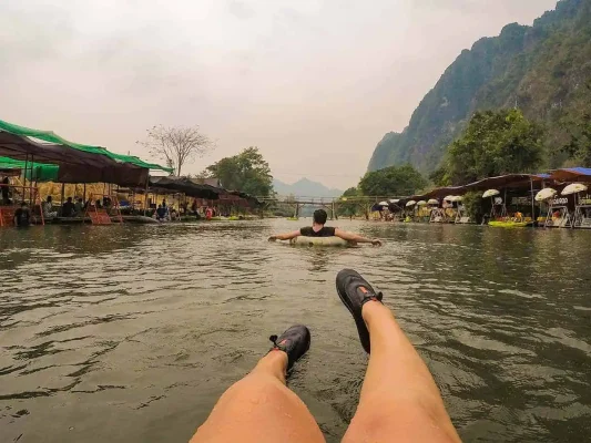 A person's legs seen from their perspective while floating on a tube in a river, following another person in the water, with a row of riverside huts and mountain scenery in the background, capturing the relaxed atmosphere of tubing in Laos.