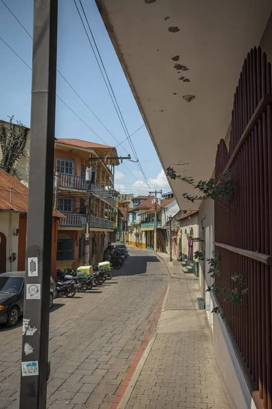 A quiet cobblestone street in a small town, lined with motorcycles and colorful buildings. The buildings feature balconies with white railings and vibrant facades. The clear blue sky provides a serene atmosphere as the street gently curves away into the distance.