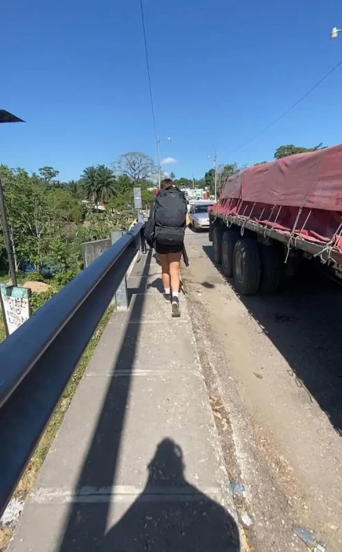 A female backpacker with a large black backpack and wearing a black tank top, shorts, and white socks walks on a narrow sidewalk beside a parked red tarp-covered truck when traveling from san ignacio to flores. The sidewalk is raised, adjacent to a guardrail overlooking a river, and leads through a small town with colorful buildings and clear skies.