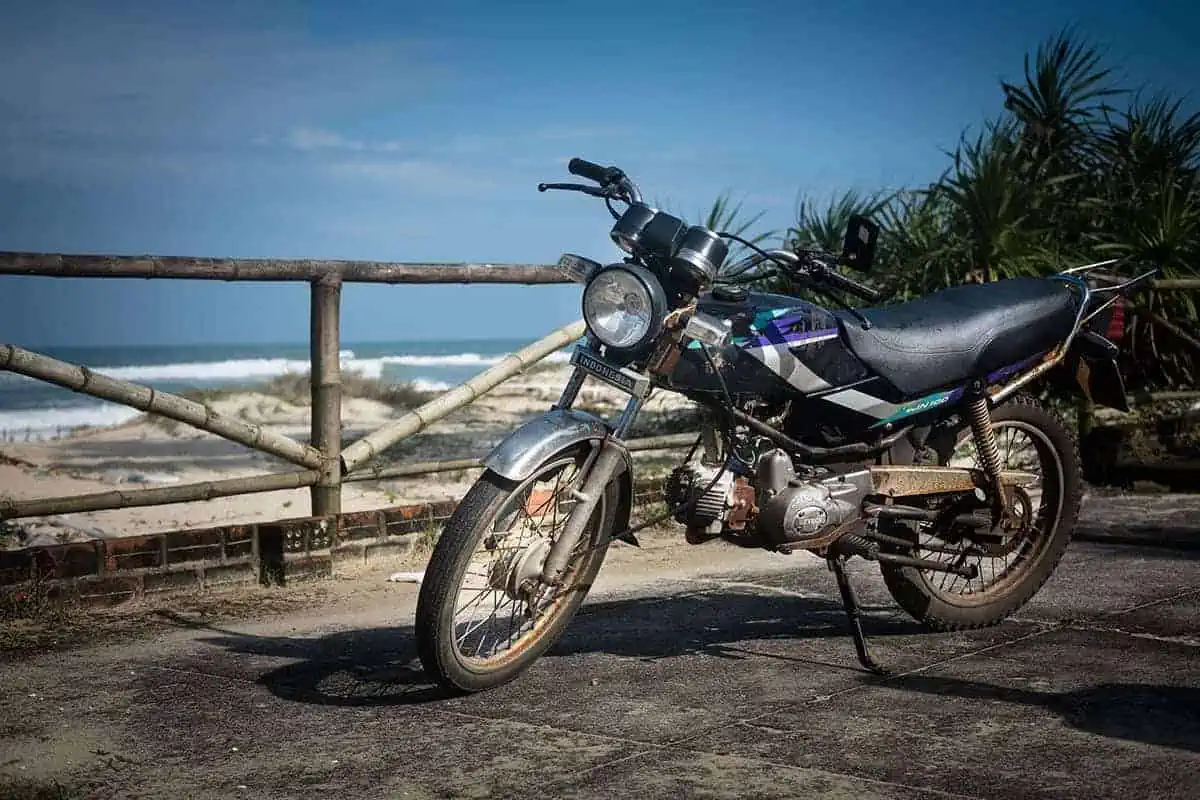 Scams In Vietnam: READ BEFORE YOU VISIT (2025) 9 A weathered motorbike parked on a concrete overlook near the beach in Vietnam, with waves crashing in the background and bamboo railing framing the ocean view.