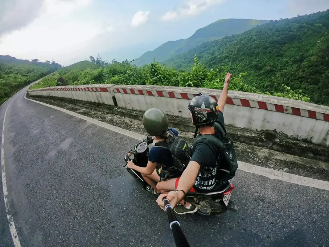 Scams In Vietnam: READ BEFORE YOU VISIT (2025) 7 Two travelers riding a motorbike on a mountain road, having avoided scams in Vietnam, with one person taking a selfie while the other steers.