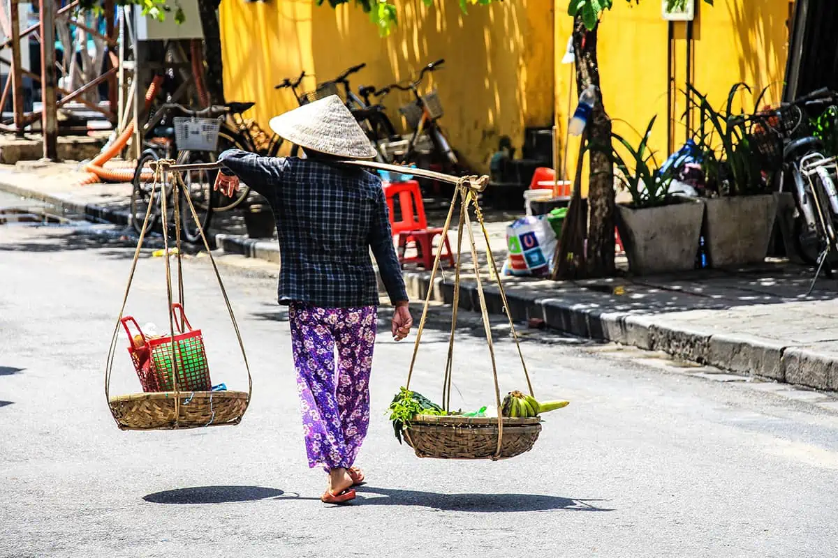 Scams In Vietnam: READ BEFORE YOU VISIT (2025) 10 A Vietnamese street vendor in a conical hat walks barefoot on the road, balancing a bamboo pole with two baskets of fresh fruit. The background features a bright yellow wall and parked bikes in Hoi An.