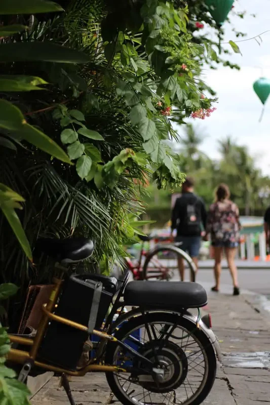 Scams In Vietnam: READ BEFORE YOU VISIT (2025) 6 A small electric bike partially hidden by greenery on a sidewalk, with two blurred pedestrians walking away in the background in Hoi An.