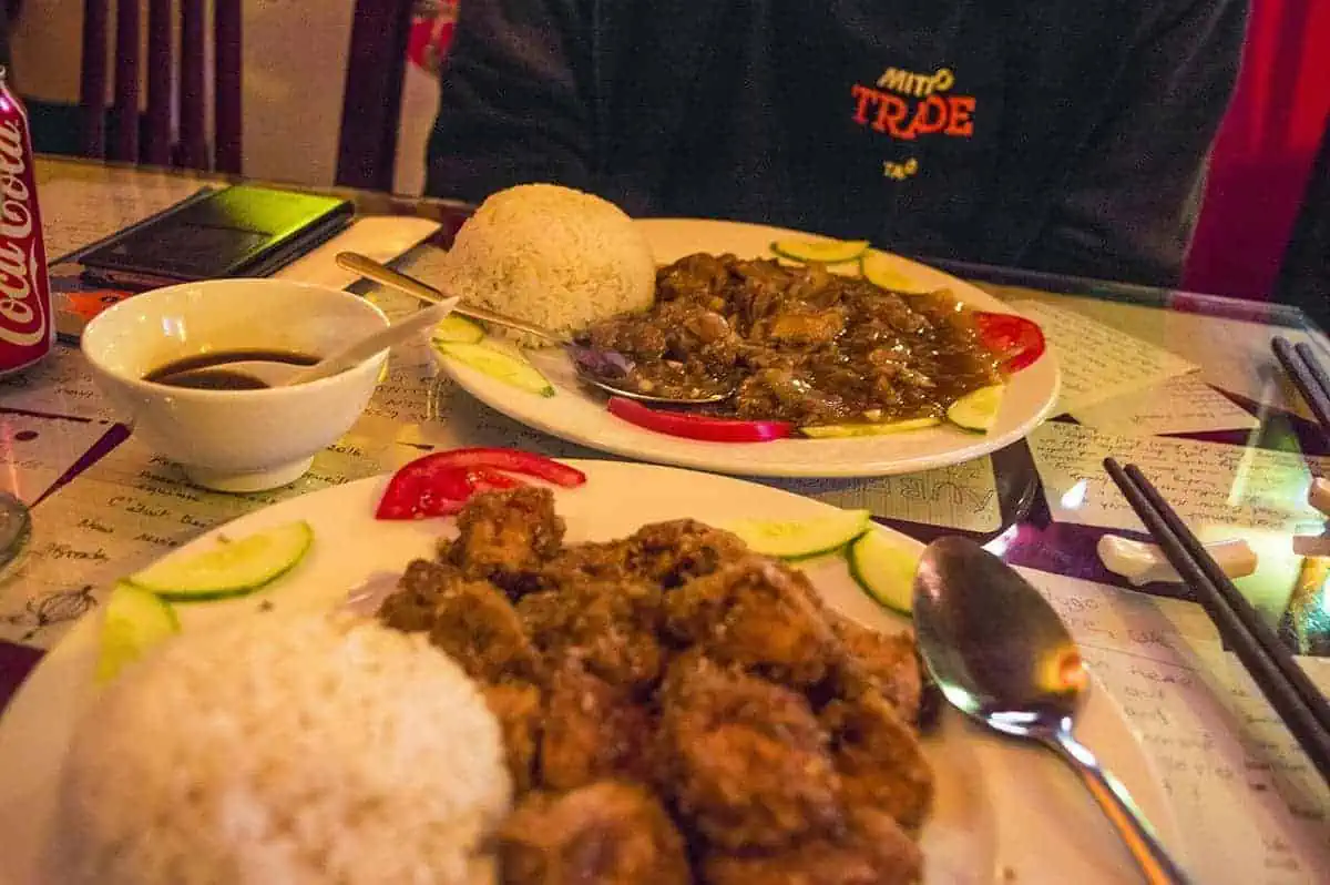 Scams In Vietnam: READ BEFORE YOU VISIT (2025) 15 Close-up of two plates of Vietnamese food featuring rice, tofu in sauce, sliced cucumbers and tomatoes, with a Coca-Cola can and soy sauce bowl on the table in Hanoi.