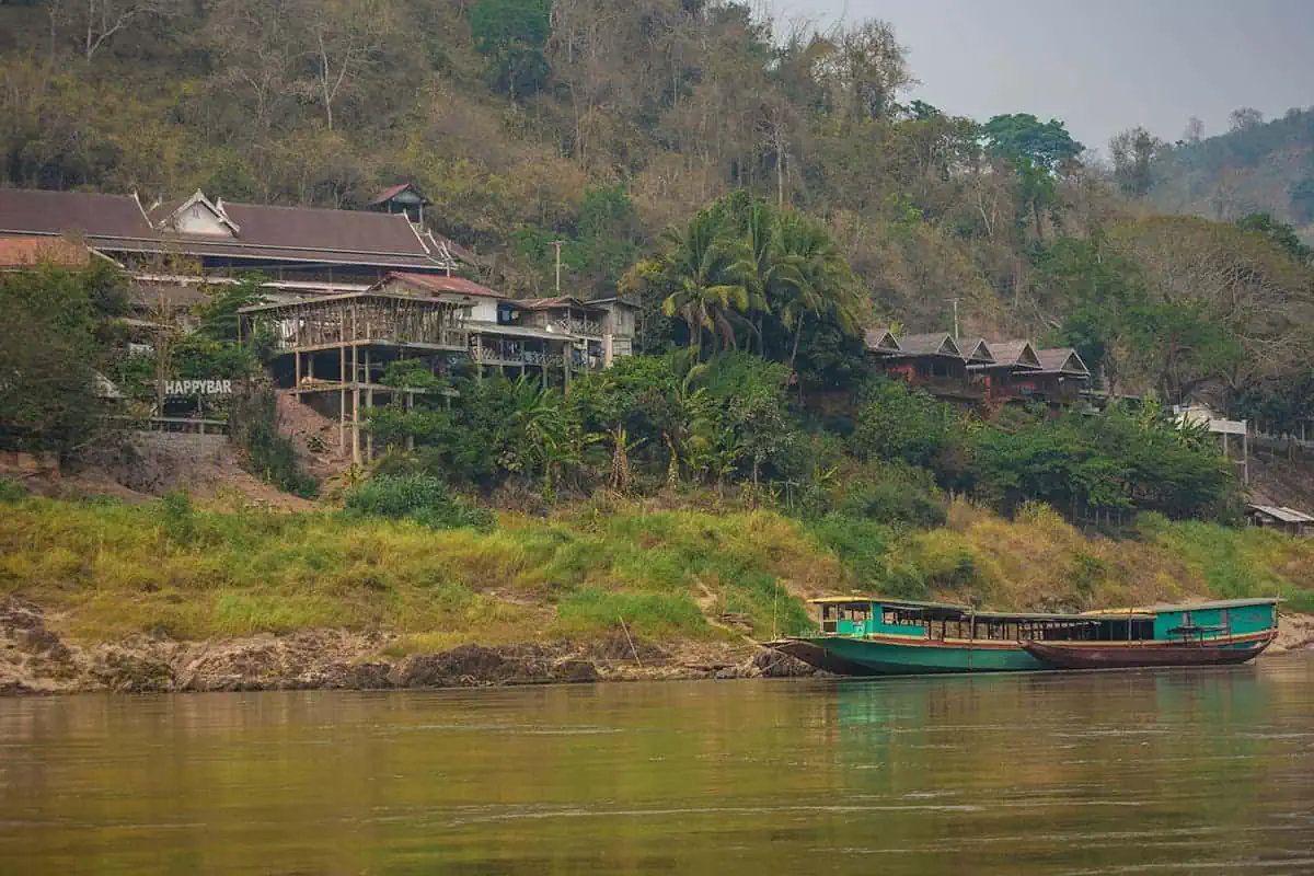 A boat cruises by a lush riverbank with traditional Lao architecture in a village called Pakbeng