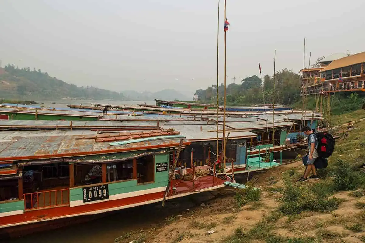 Travelers with backpacks approach a fleet of slow boats docked at the riverbank