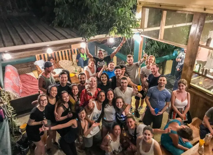 A large group of smiling people, gathered closely together for a group photo at night. The scene is set in a cozy, well-lit outdoor area with trees and string lights creating a warm ambiance. Everyone appears to be enjoying themselves, holding drinks and posing for the camera. This image highlights the communal and festive experiences often encountered during solo travel in Belize.