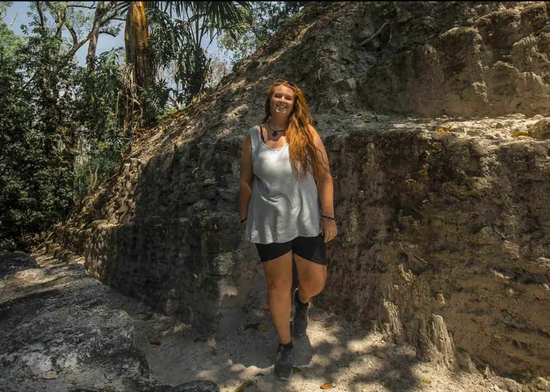 tasha amydressed in casual shorts and a tank top, stands on an ancient stone pathway surrounded by dense jungle vegetation. She leans slightly against a large stone structure, with sun-dappled light filtering through the trees above. Her expression is one of joy and contentment, capturing the essence of discovering historical and archaeological wonders in Belize.