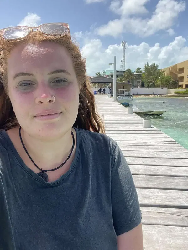A close-up selfie of tasha amy with auburn hair, wearing a grey T-shirt and sunglasses perched on her head, standing on a long wooden pier that stretches into the turquoise sea. The background features a mix of beachfront buildings and swaying palm trees under a bright sky. Her slight smile and sun-kissed face reflect the joy and relaxation of solo travel in Belize.