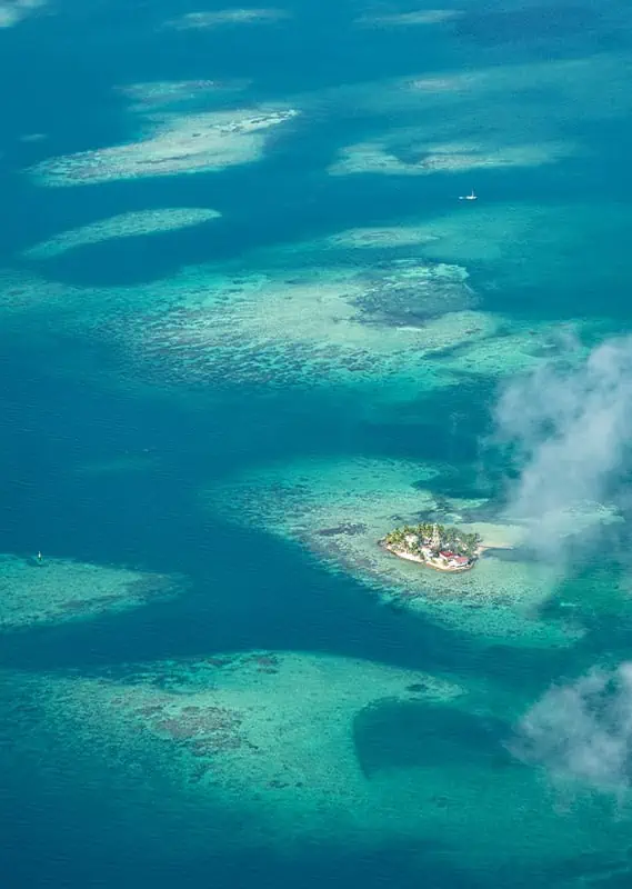 An aerial view of a tiny, lush tropical island surrounded by vibrant turquoise waters and intricate coral reefs. The island is dotted with a few palm trees and a small building, creating a picturesque and serene landscape. Wisps of clouds cast shadows on the water, adding depth to the scene. This image epitomizes the secluded beauty and tranquil experiences awaiting travelers in Belize.