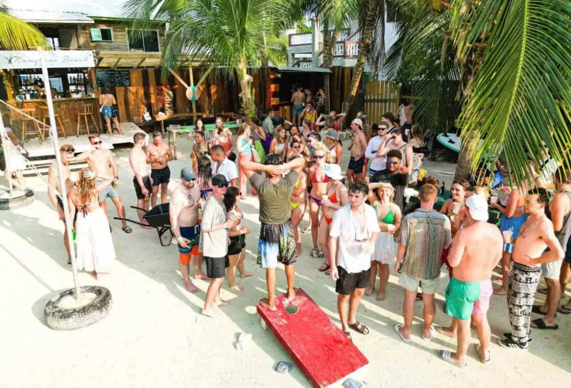A lively beach party scene with numerous people gathered around a sandy area in front of a rustic beach bar. The crowd, dressed in swimwear, is engaged in conversations and activities under the shade of palm trees. The vibrant atmosphere showcases the social side of solo travel in Belize, where travelers can meet and enjoy the company of others in a tropical setting.