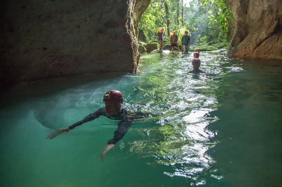 Solo Travel In Belize: My Experience As A Solo Female Backpacker 9 A group of people, equipped with helmets and headlamps, explore an emerald green water-filled cave. In the foreground, a person swims towards the camera, stretching their arms forward. The background reveals a small group standing on rocks at the cave's mouth, bathed in natural light filtering through the lush jungle foliage. This scene captures the thrill and beauty of solo travel in Belize, showcasing the adventure of exploring natural cave systems.
