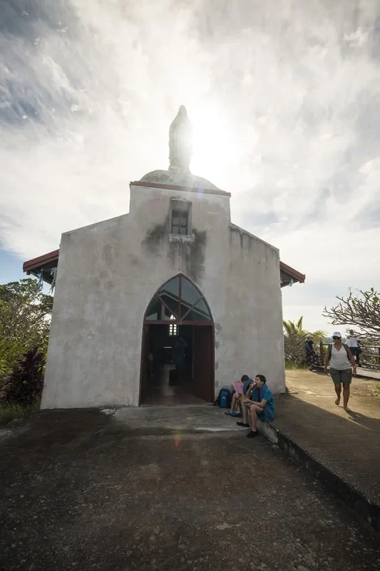 14 BEST Things To Do In Lifou: Cruise Passengers & More 6 A quaint chapel with a statue on top bathed in sunlight, with people sitting and walking around, a sight to explore in Lifou from a cruise ship.
