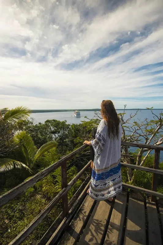 14 BEST Things To Do In Lifou: Cruise Passengers & More 7 tasha amy standing on a wooden walkway overlooking the ocean, with a cruise ship in the distance, highlighting the peaceful ambiance and things to do in Lifou.