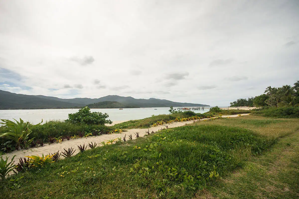 Looking across a grassy patch and sandy trail on Mystery Island toward a wooden pier and the mountainous coastline across the water.