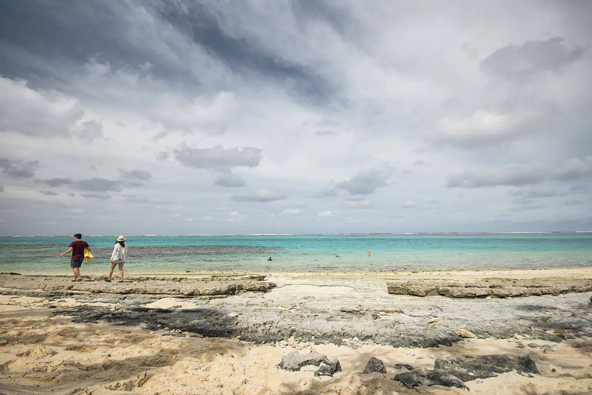 Two people walk along the rocky beach of Mystery Island under a dramatic, cloud-filled sky, while others swim in the shallow turquoise lagoon.