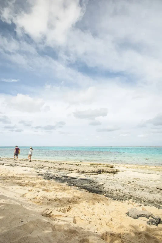 A couple walks along the edge of Mystery Island’s sandy and rocky beach, with clear blue waters and a horizon that meets the sky.