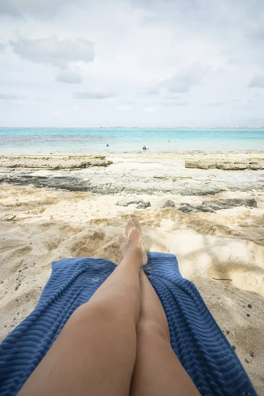 Close-up of tasha amy’s legs resting on a blue towel on the sand of Mystery Island, facing the calm ocean and coral-strewn shoreline.