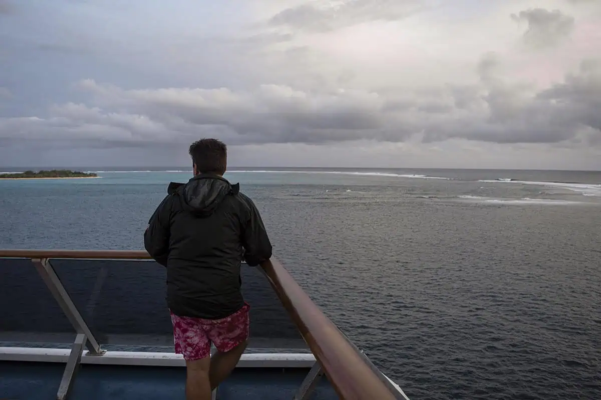 A man in a black jacket and red swim trunks stands on a cruise ship deck, gazing at Mystery Island across turquoise waters under a moody, overcast sky.