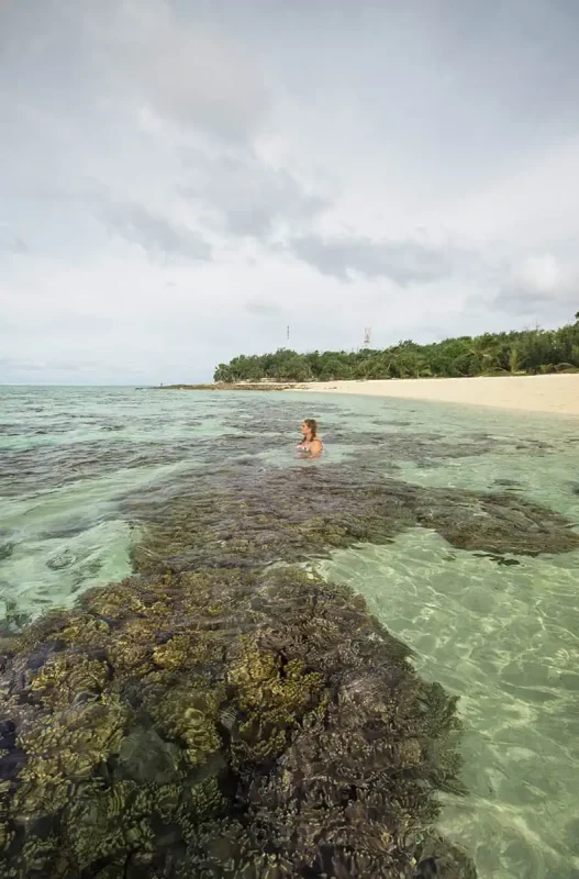 tasha amy stands waist-deep in the clear lagoon of Mystery Island, surrounded by coral heads and facing a peaceful, undeveloped beach with forested hills beyond.