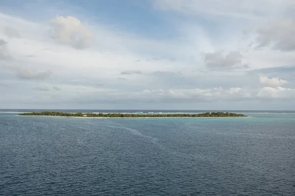 A wide aerial view of Mystery Island shows its thin strip of green vegetation surrounded by turquoise shallows and deep blue ocean under a partly cloudy sky. Come check out the best things to do on mystery island for cruise passengers
