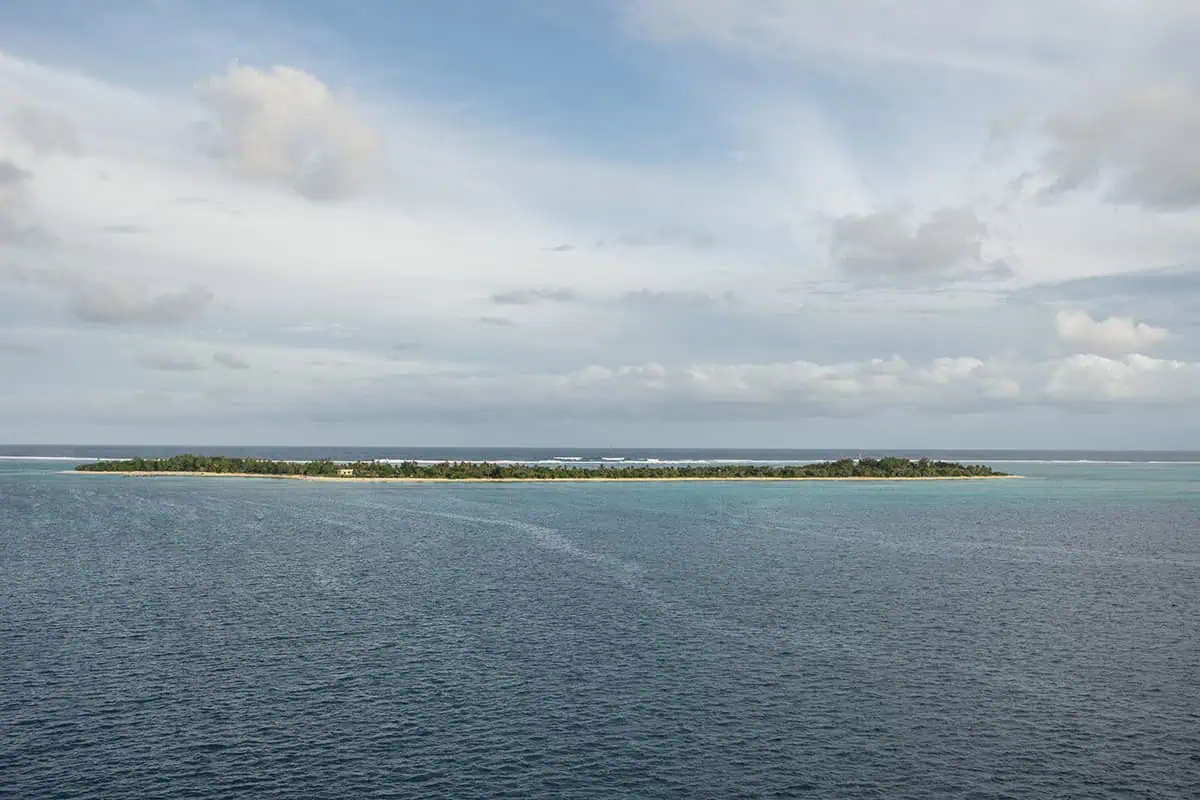 A wide aerial view of Mystery Island shows its thin strip of green vegetation surrounded by turquoise shallows and deep blue ocean under a partly cloudy sky. Come check out the best things to do on mystery island for cruise passengers