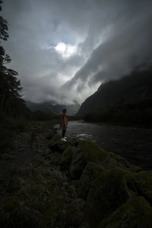 tasha amy n a raincoat stands on mossy rocks beside a river, gazing at the mist and cliffs of Milford Sound.