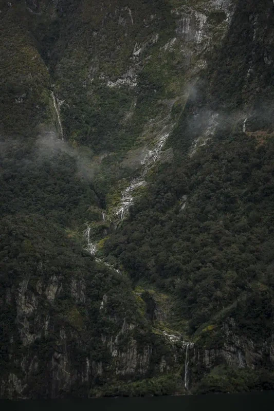 Multiple thin waterfalls stream down the face of a steep mountain in Milford Sound.