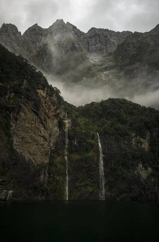 Twin waterfalls called fairy falls descend from a misty cliff into the deep waters of Milford Sound, with mountains in the background.