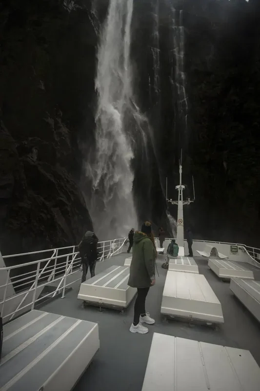 Visitors on the upper deck of a cruise boat stand in awe beneath a powerful stirling falls crashing down directly in front of them in Milford Sound.