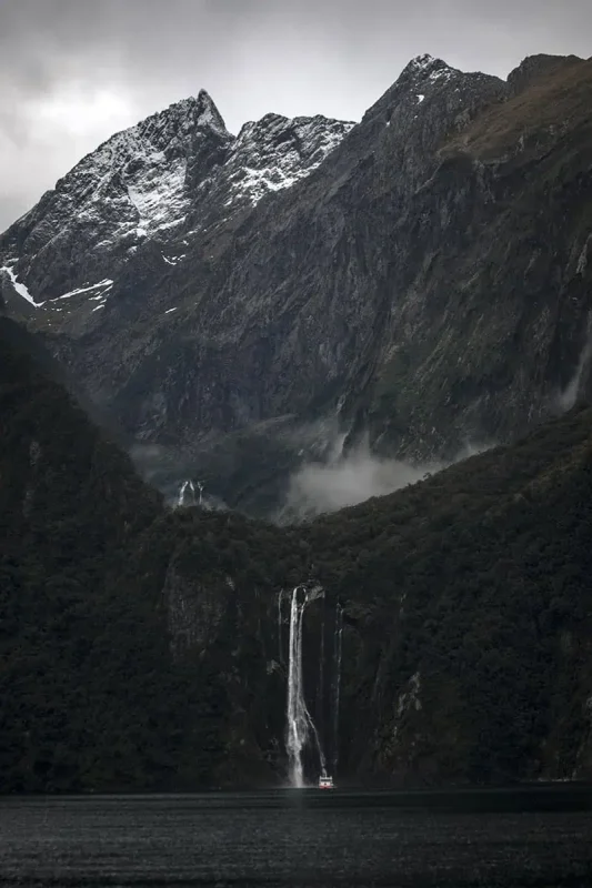 A dramatic waterfall plunges from a forest into the fiord below, with a small cruise boat at its base and snow-dusted peaks behind.
