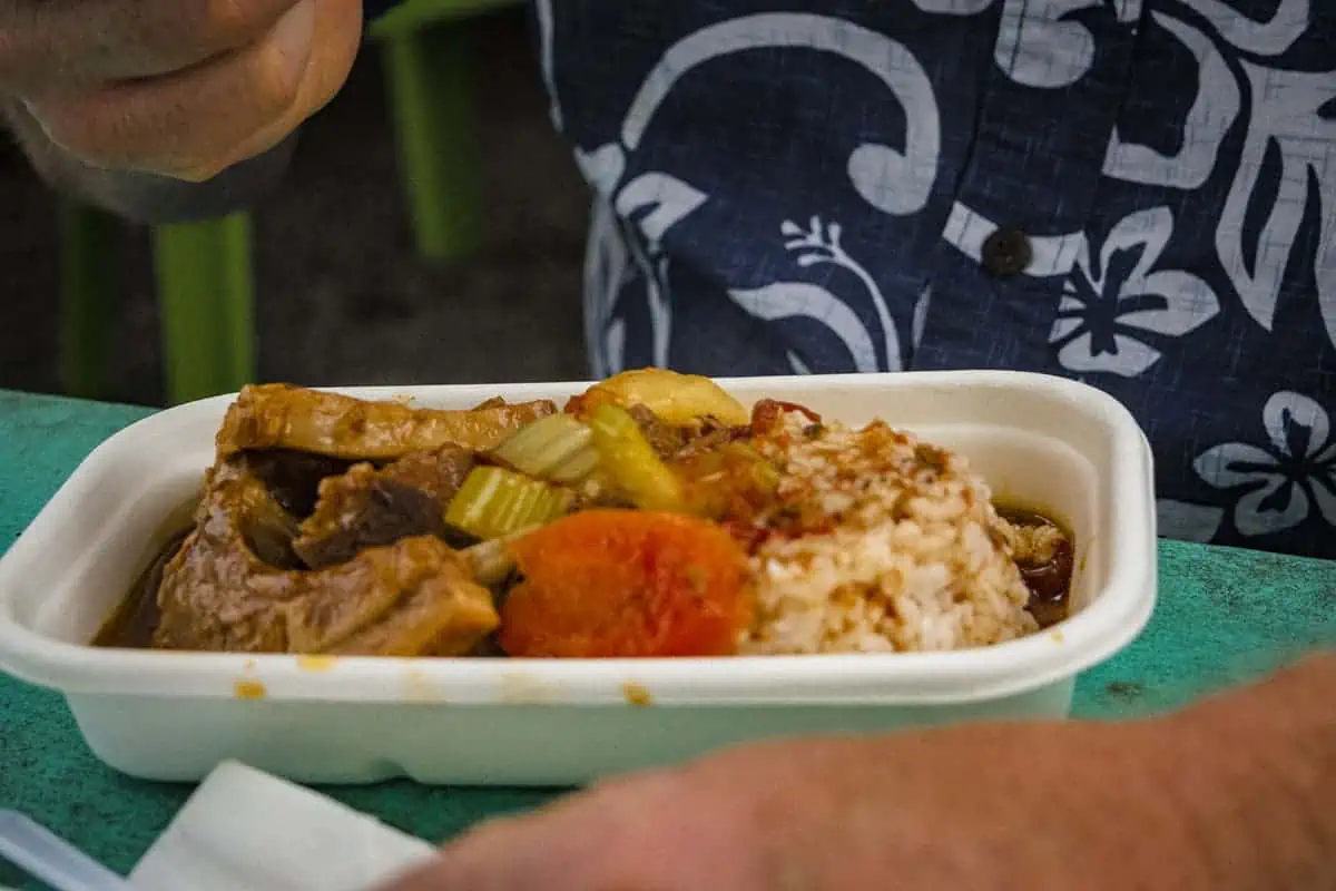 Where To Eat In Rarotonga: BEST Restaurants & Cafes 2025 21 A hearty meal of beef stew and rice in a takeout container, a local delicacy from the Muri Night Market, a popular place to eat in Rarotonga, held by a person in a floral shirt.