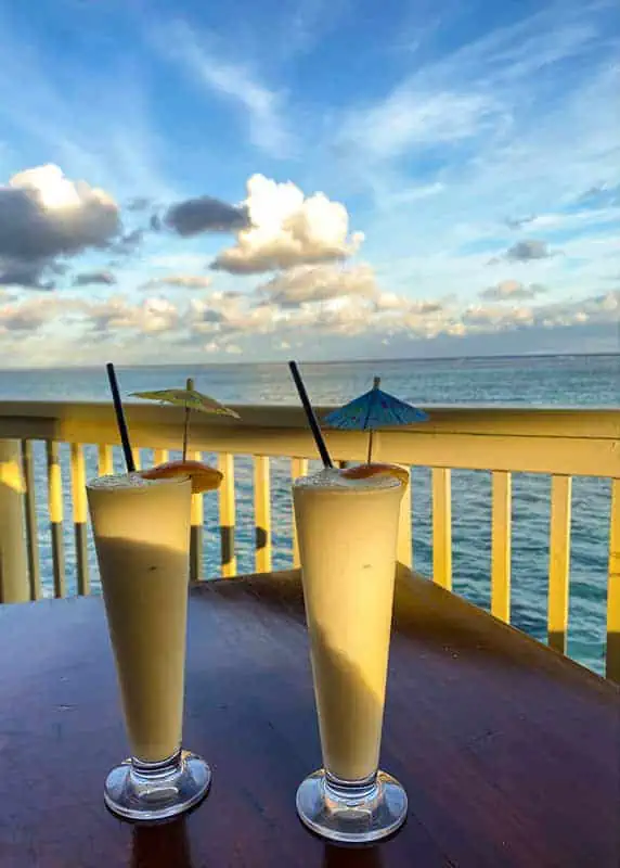 Where To Eat In Rarotonga: BEST Restaurants & Cafes 2025 27 Two creamy pina coladas with decorative umbrellas against a seaside backdrop, served at captain andys bar and grill at the rarotongan