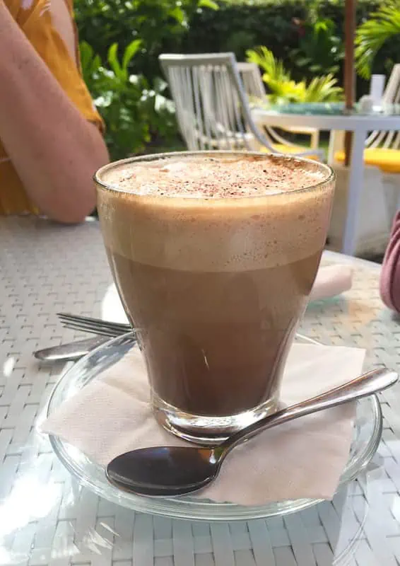Where To Eat In Rarotonga: BEST Restaurants & Cafes 2025 14 Frothy cappuccino in a glass cup, a morning favorite at the Lazy Trout among the best cafes in Rarotonga, with a serene garden backdrop.