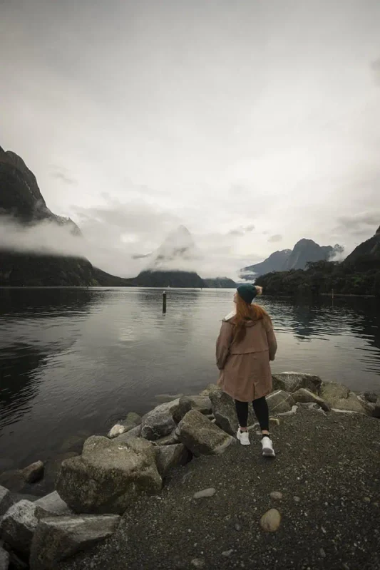Milford Sound In Winter: Is It Still Worth The Hype? 15 tasha amy stands on a rocky outcrop by the water's edge, wearing a tan coat and dark beanie. The calm waters of Milford Sound reflect the mist-covered peaks in the distance, creating a serene and contemplative winter scene in Milford Sound.
