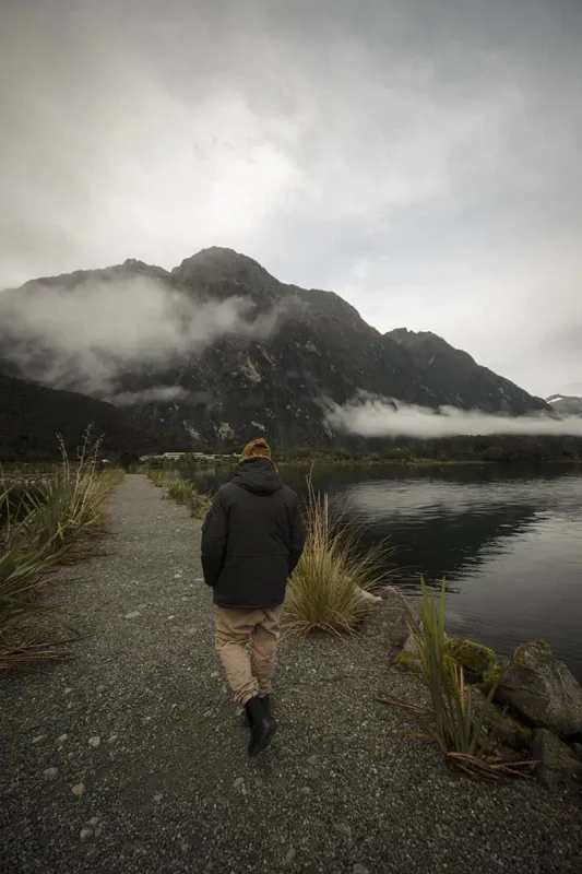 Milford Sound In Winter: Is It Still Worth The Hype? 11 A person in a dark coat and mustard-yellow beanie walks along a gravel path beside a calm, misty lake. The towering, cloud-covered mountains in the background create a dramatic and serene atmosphere, typical of winter in Milford Sound.