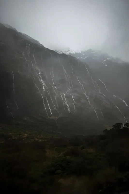 Milford Sound In Winter: Is It Still Worth The Hype? 8 Steep, rugged mountains draped in mist and rain, with multiple thin waterfalls cascading down their slopes. The scene is dramatic and moody, highlighting the raw natural beauty and frequent rains typical of Milford Sound's winter season.