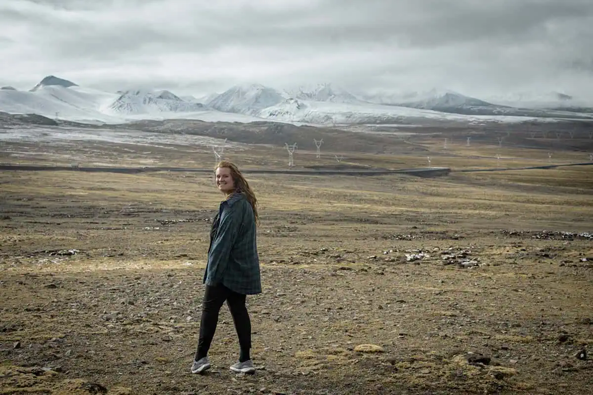 120+ Would You Rather Travel Questions For Ultimate Wanderers 1 tasha amy in a green flannel shirt stands in a windswept Tibetan landscape, with dramatic, snow-covered peaks rising in the distance