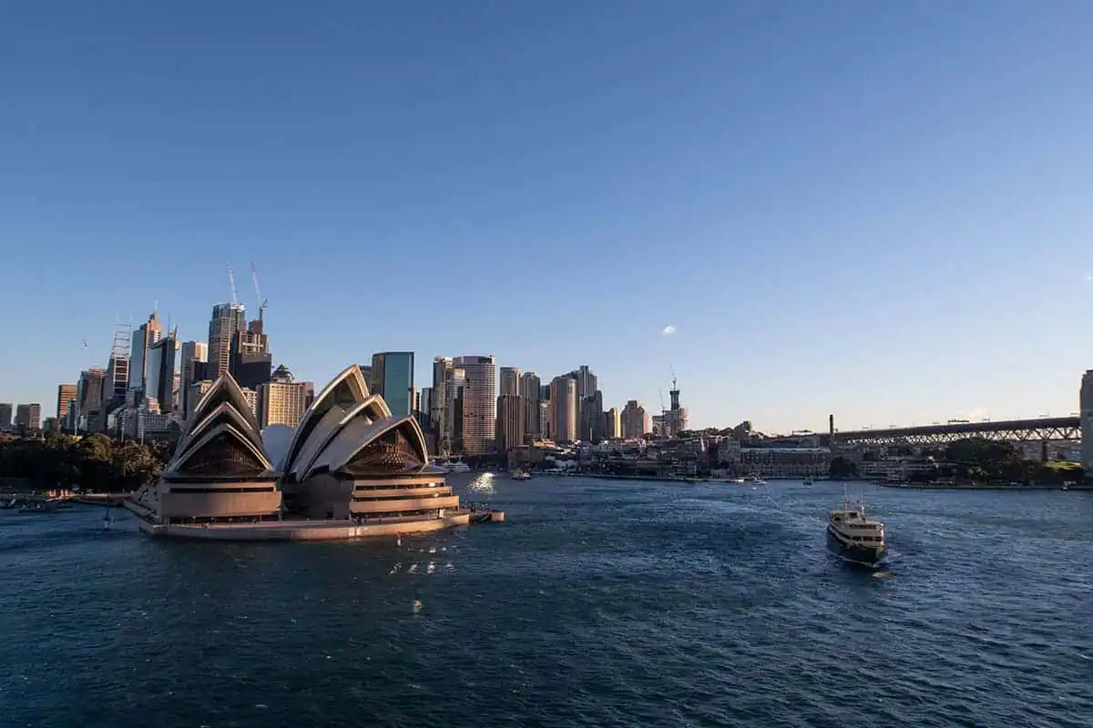 120+ Would You Rather Travel Questions For Ultimate Wanderers 6 The Sydney Opera House with a ferry in the water, tall buildings and a clear sky.