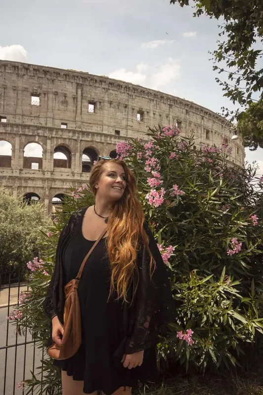 120+ Would You Rather Travel Questions For Ultimate Wanderers 8 tasha amy in black stands near oleander flowers, with the Roman Colosseum behind her.