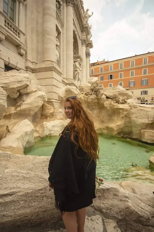 120+ Would You Rather Travel Questions For Ultimate Wanderers 9 tasha amy poses next to the iconic Trevi Fountain, surrounded by intricate sculptures and pale blue water with historic buildings in the background.
