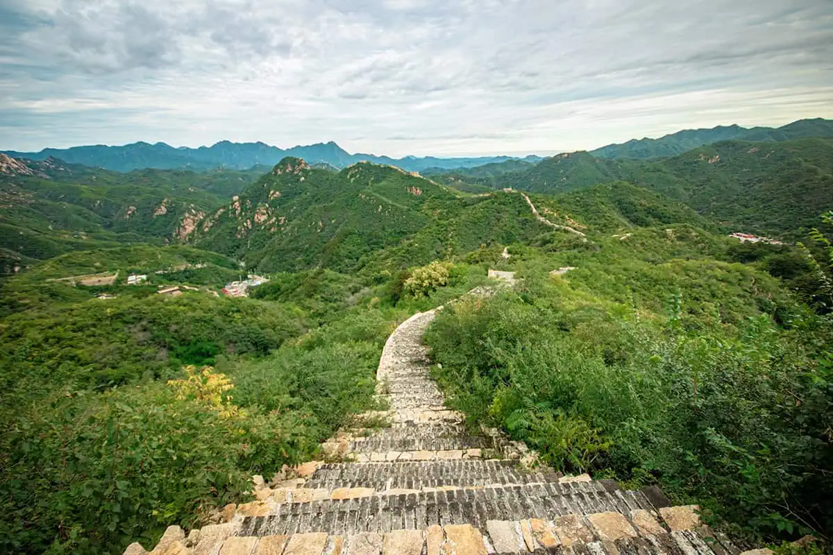 A rugged section of the Great Wall of China stretches through green hills, with distant mountain peaks, this location should definitely be considered for the would you rather travel questions